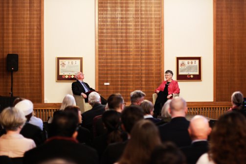 Professor John Naughton and Professor Jane Clarke, the President of Wolfson, sit at the front of the room in front of a crowd of people.