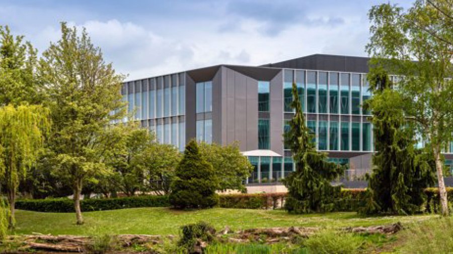 Modern office building with large glass windows surrounded by lush greenery and trees under a cloudy sky.