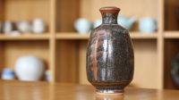 A dark brown vase with orange symbol on in front of a blurred background of more pottery on shelves.