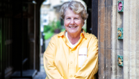 Photograph of Sandi Toksvig, wearing a yellow shirt and leaning against a stone entryway.