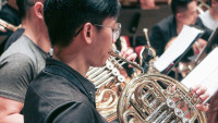  A man plays a french horn in an orchestra, surrounded by other musicians with sheet music stands visible.